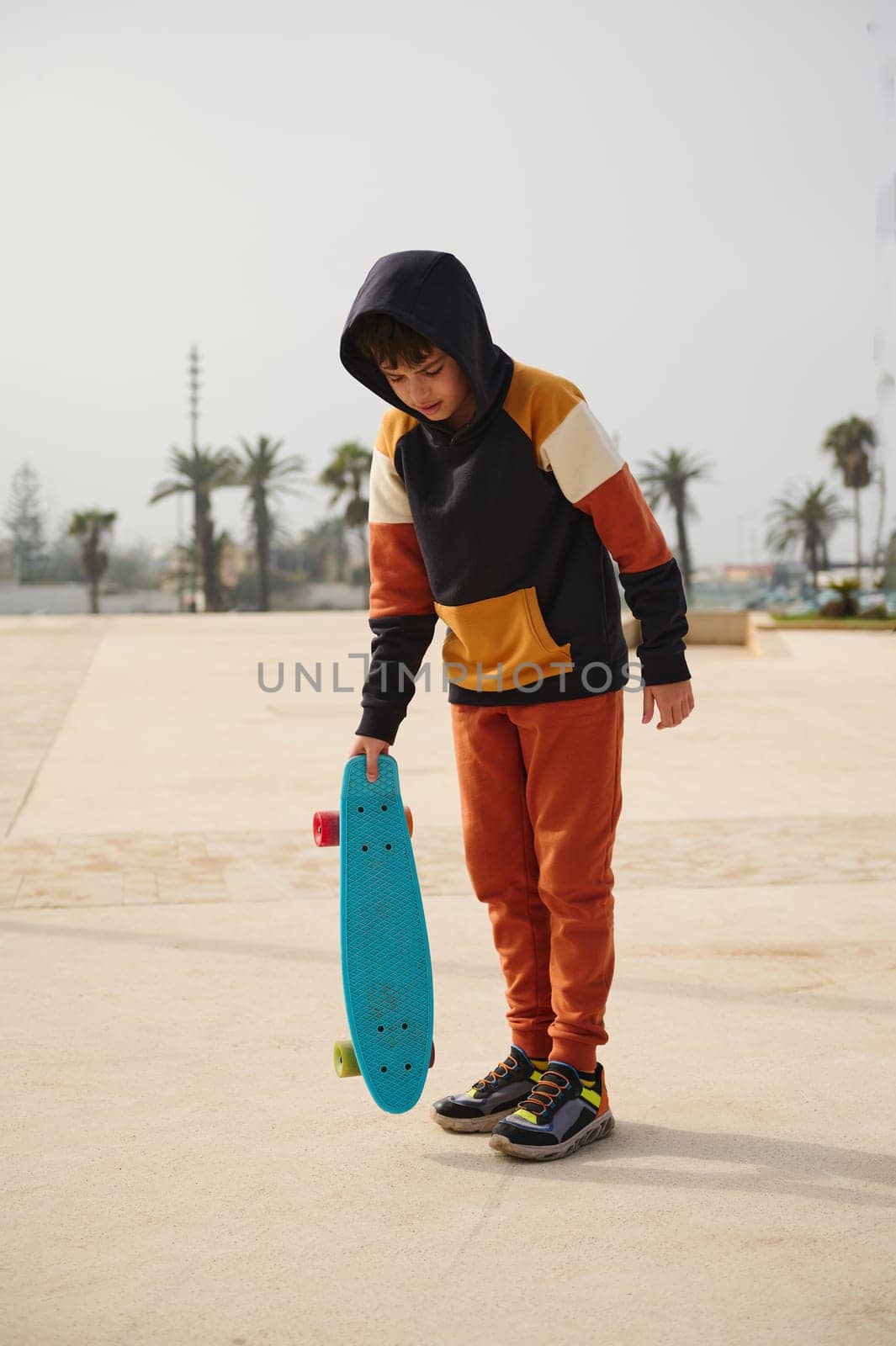 Vertical full length portrait of a multi ethnic teenager, stylish schoolboy in sportswear, holding skateboard, standing on an outdoor skatepark playground. People. Childhood and extreme sport concept