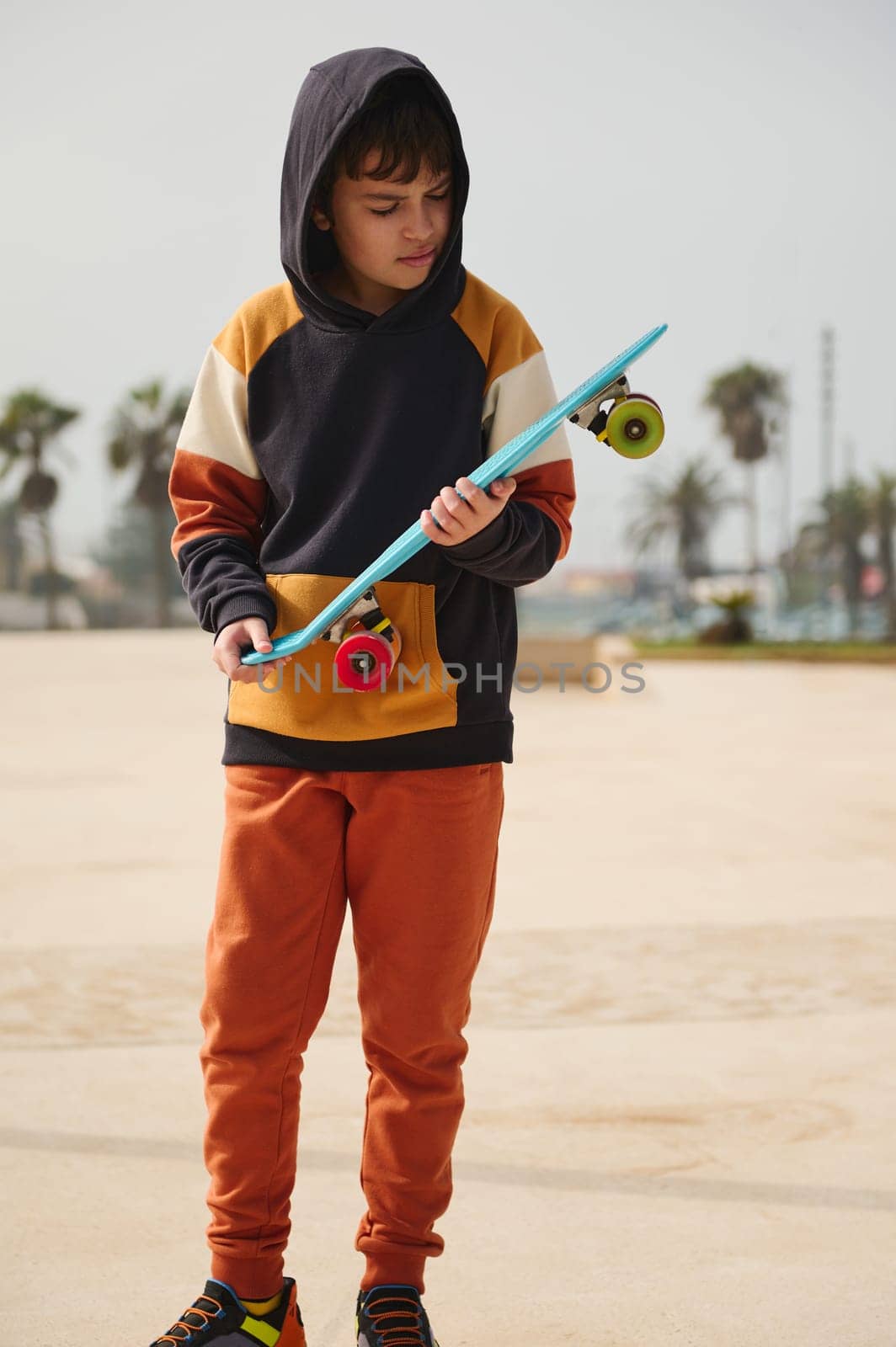 Adorable active Caucasian teenage boy dressed in stylish sportswear with hood in head, holding skateboard and looking down, standing on an outdoor skatepark, enjoying weekend activities outdoors