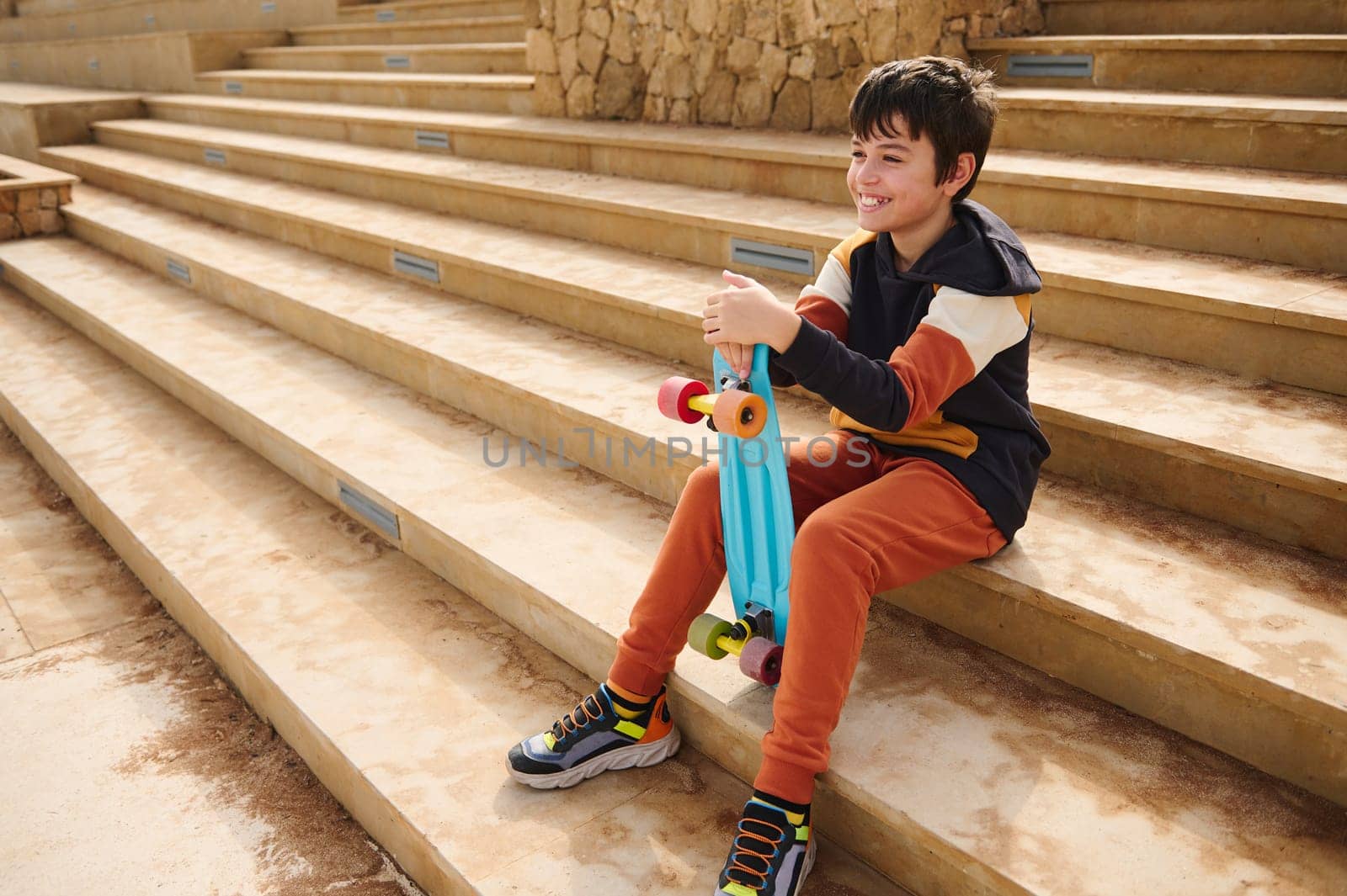 Full length horizontal portrait of multi ethnic teenager boy in activewear, holding his blue skateboard, smiling looking away, sitting on steps outdoors. People. Sport. Copy advertising space for text