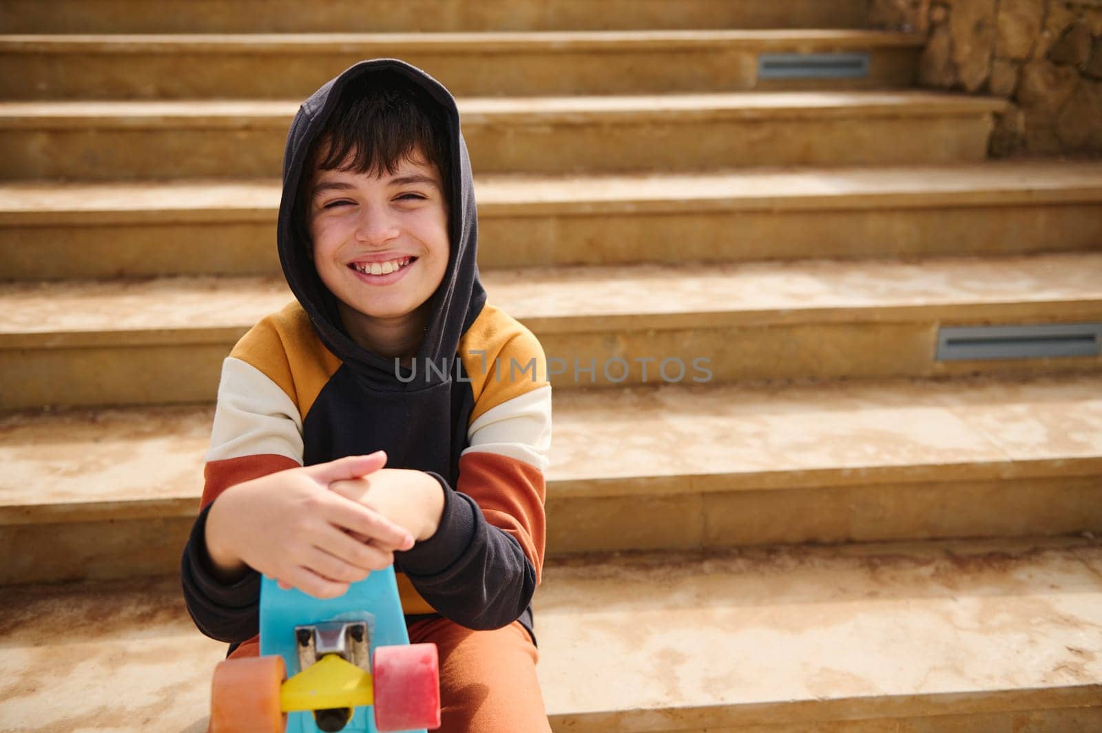 Authentic portrait of charming cheerful multi ethnic teenage boy, hipster holding skateboard, cutely smiles looking at camera, sitting on marble steps, dressed in stylish colorful trendy hoodie