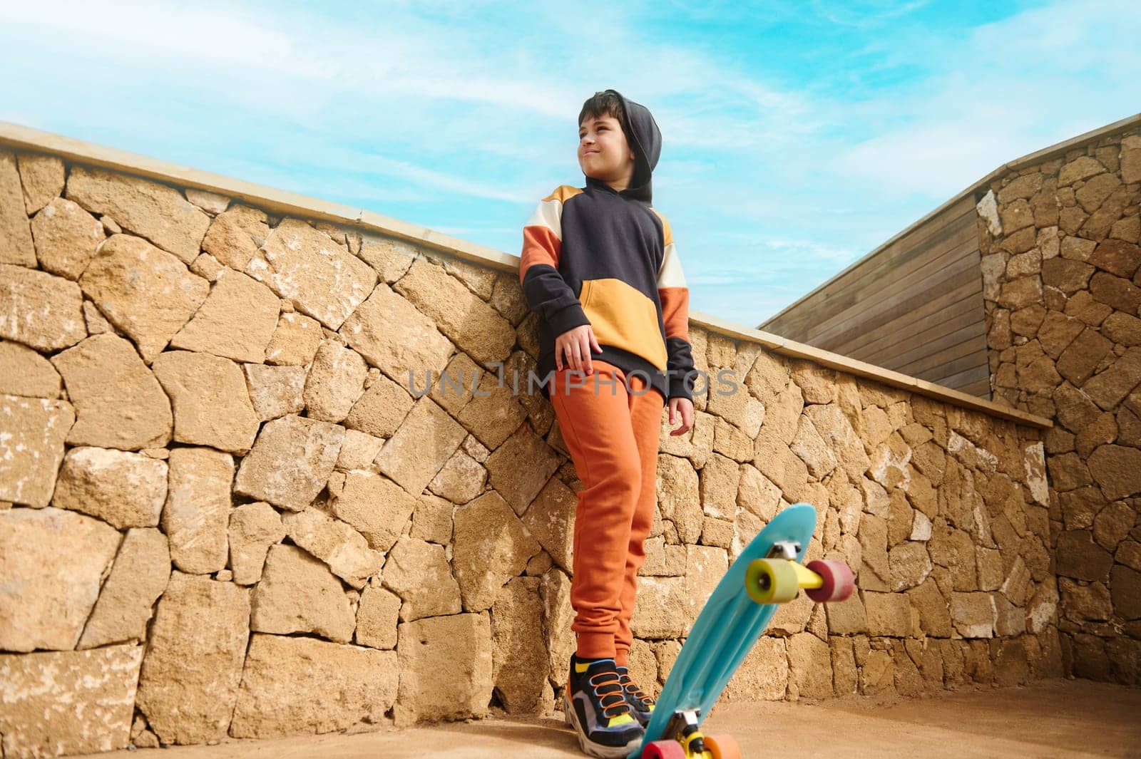 Full length portrait of handsome teenager boy in sportswear, playing with skateboard, dreamily looking aside, standing outdoors against a stone wall background. People. Sport. Active healthy lifestyle