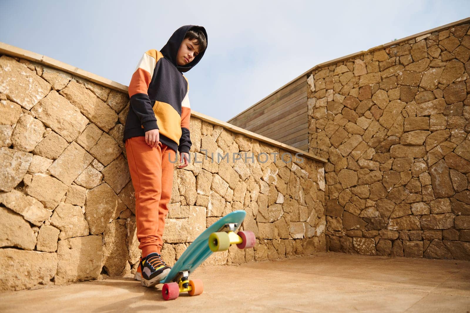 Full length portrait of handsome teenager boy in sportswear, playing with a skateboard, standing outdoors with hands in pockets against a stone wall background. People. Sport. Active healthy lifestyle