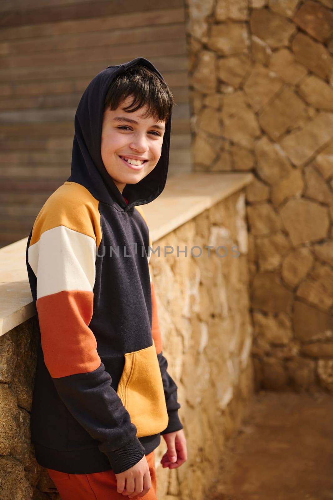 Portrait of multi ethnic happy cheerful smiling teenager boy hipster dressed in multi colored stripped sweatshirt hoodie, looking at camera, standing against a stone wall background. Copy ad space