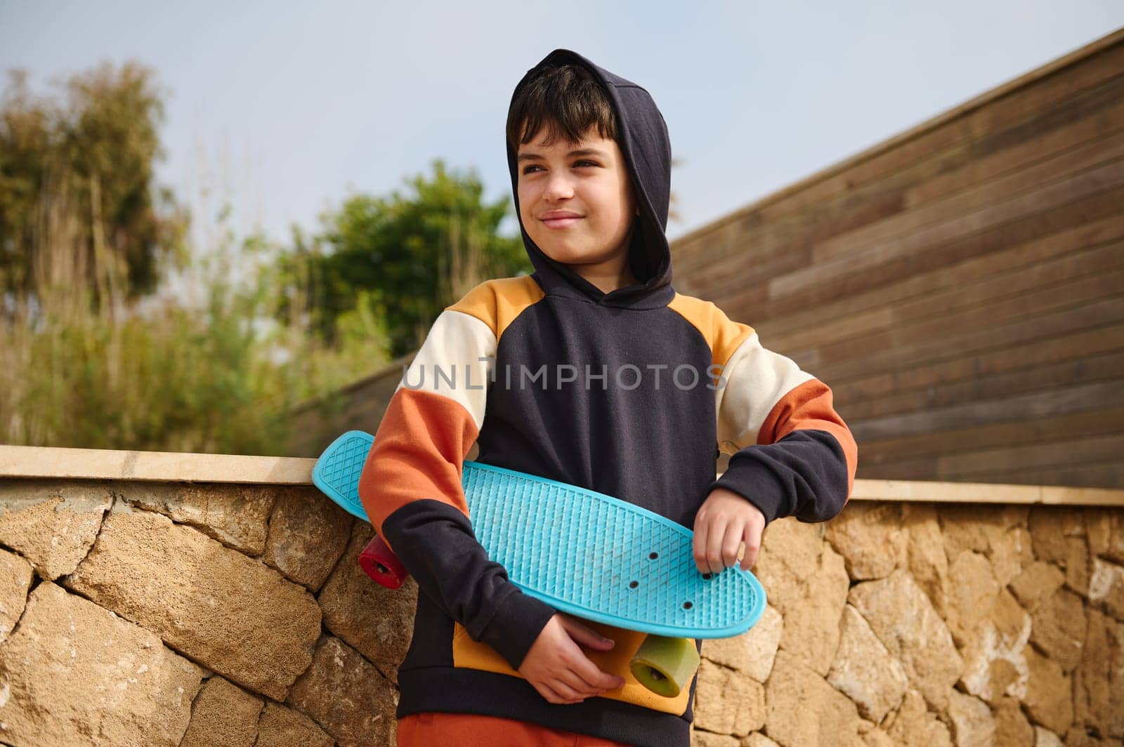 Lifestyle portrait of a teenager boy, hipster, skateboarder in trendy hoodie, holding his blue skateboard, smiling looking away, standing outdoors, against a stone wall background. Copy ad space