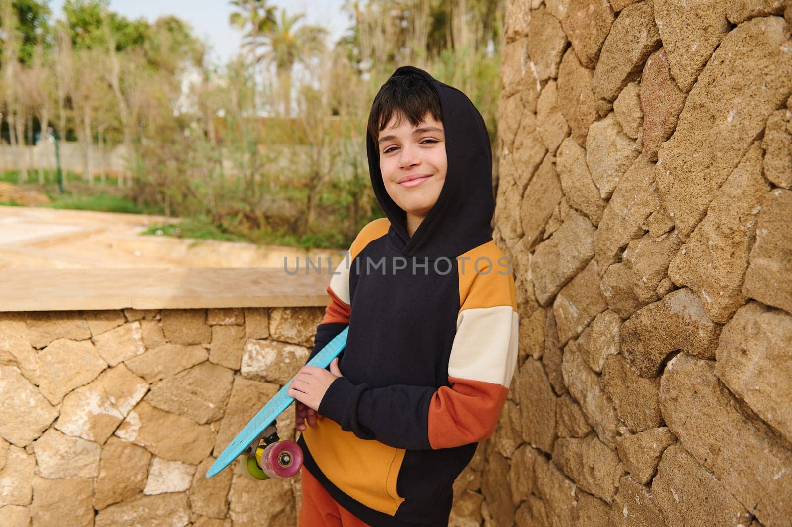 Authentic portrait of charming Caucasian teenage boy, holding skateboard, cutely smiles looking at camera, standing against beige stone wall background, dressed in black sweatshirt. Extreme sport