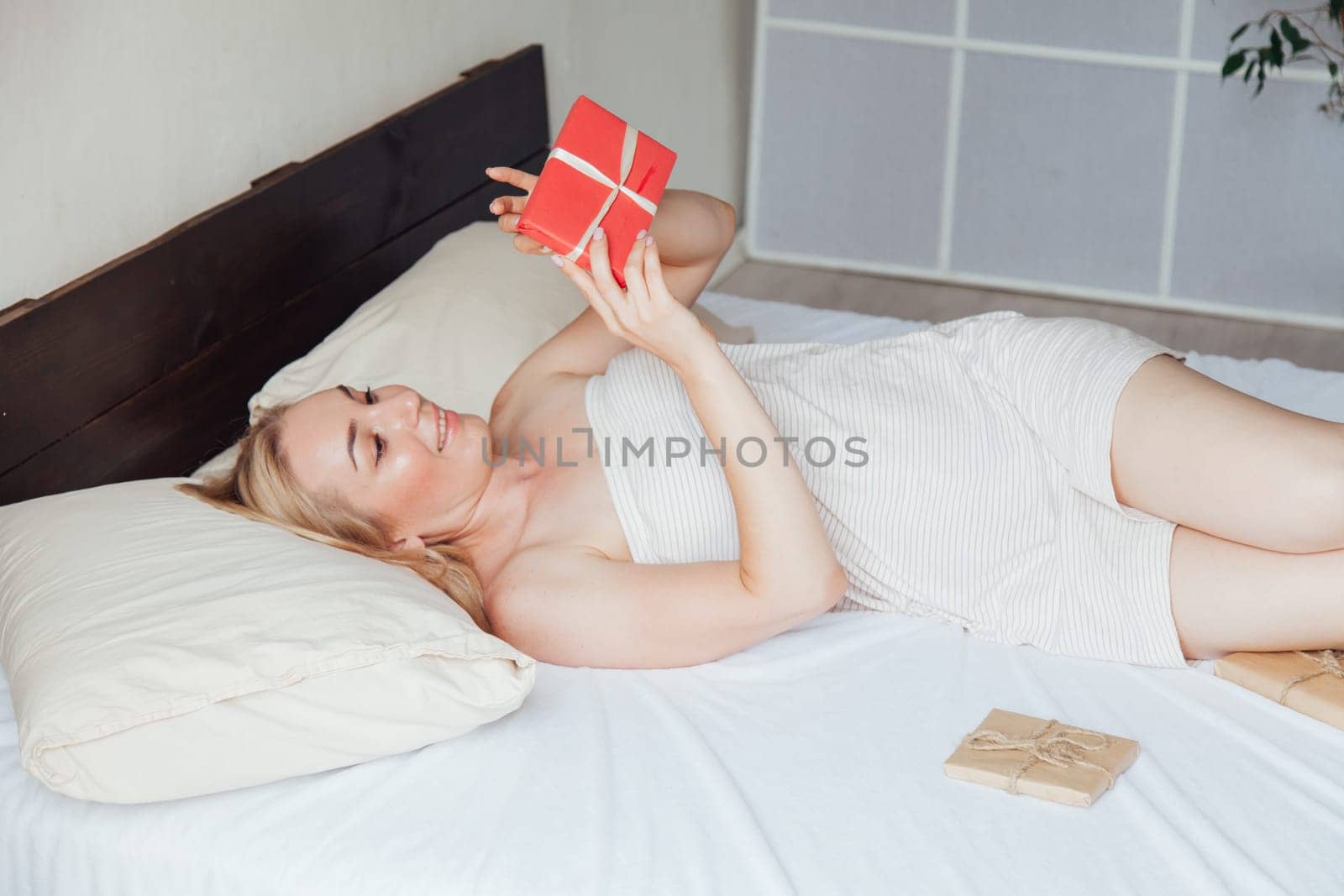 Beautiful blonde woman opens holiday gift in bedroom in bed by Simakov
