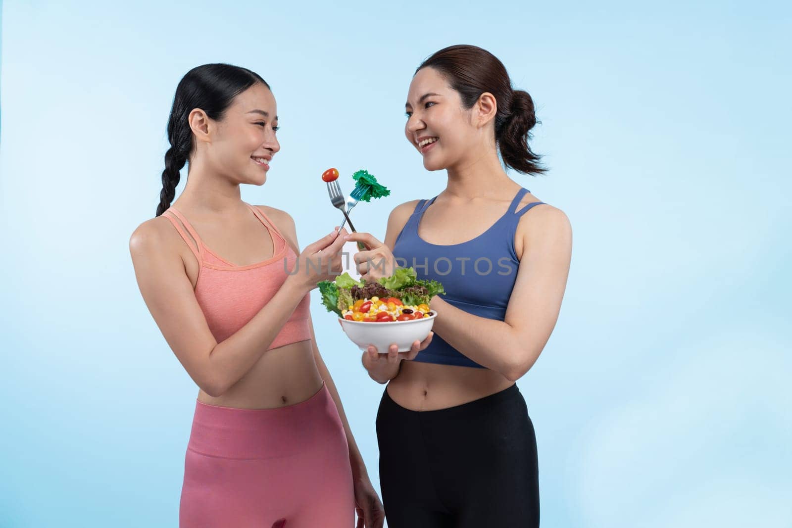 Two young sporty Asian women in sportswear holding salad bowl fill with fruit and vegetable. Natural youthful and fit body lifestyle people with balance nutrition on isolated background. Vigorous