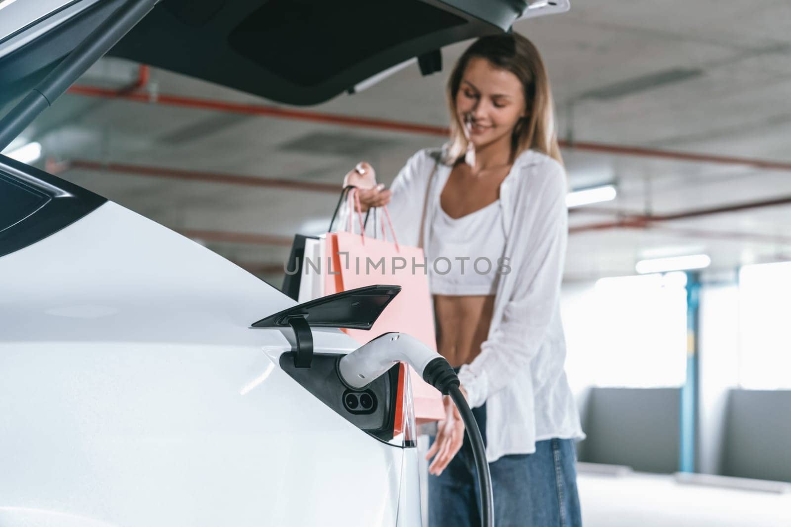 Young woman travel with EV electric car to shopping center parking lot charging in downtown city showing urban sustainability lifestyle by green clean rechargeable energy of electric vehicle innards