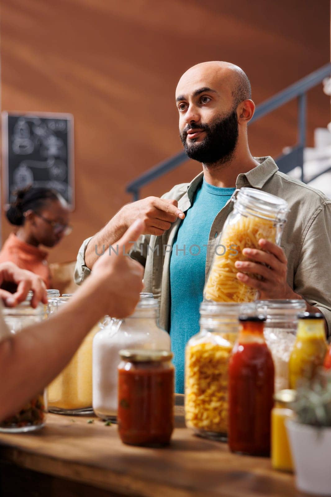 Middle Eastern customer holds and points at glass jar filled with a certain organic food product while another man gestures thumbs up in grocery store. Also in store was a black woman looking around.