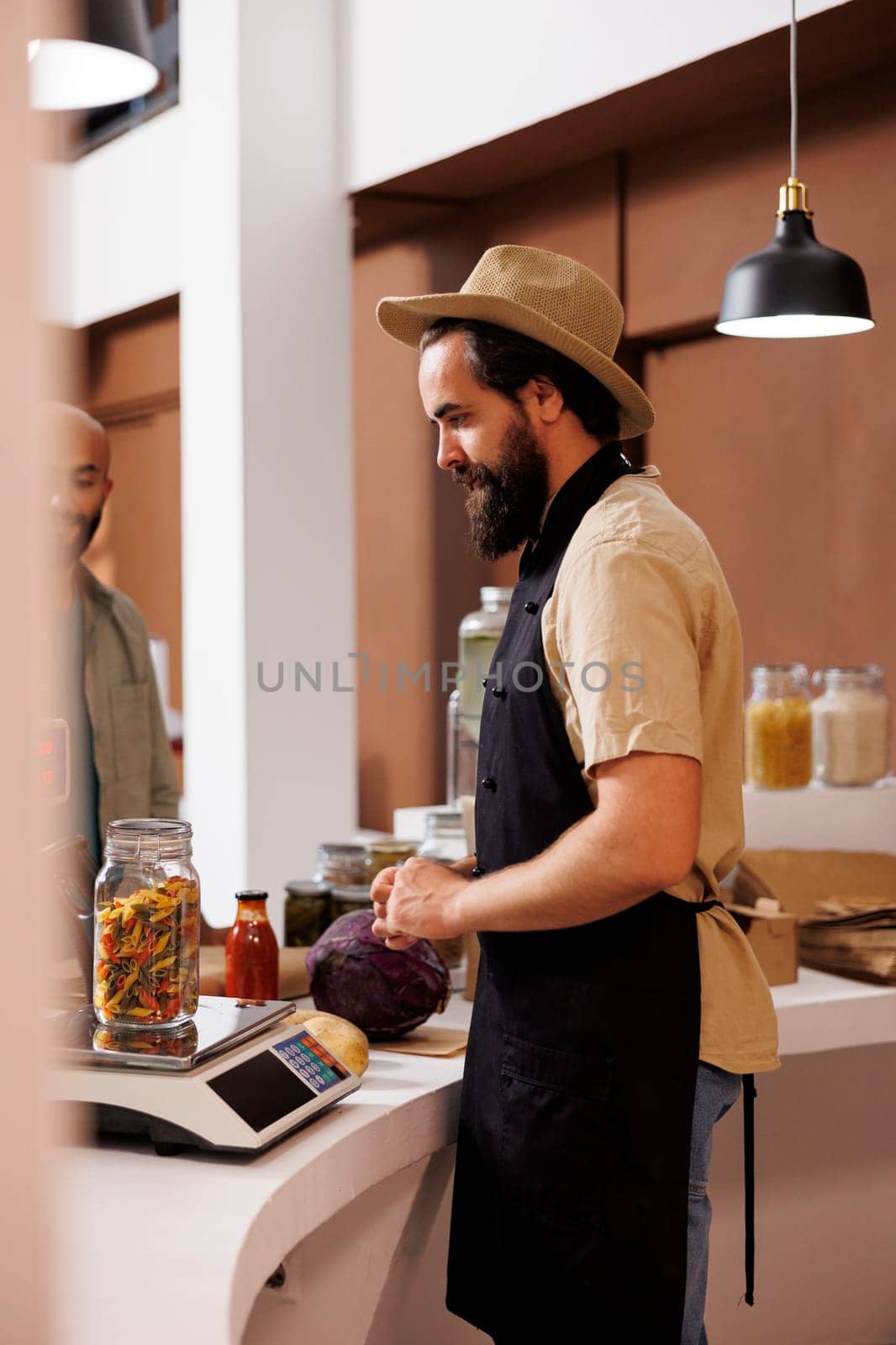 Male storekeeper, wearing a hat and apron, stands behind cashier desk while looking at jar filled with certain organic food item on measuring scale. Caucasian vendor weighing pasta for customer.