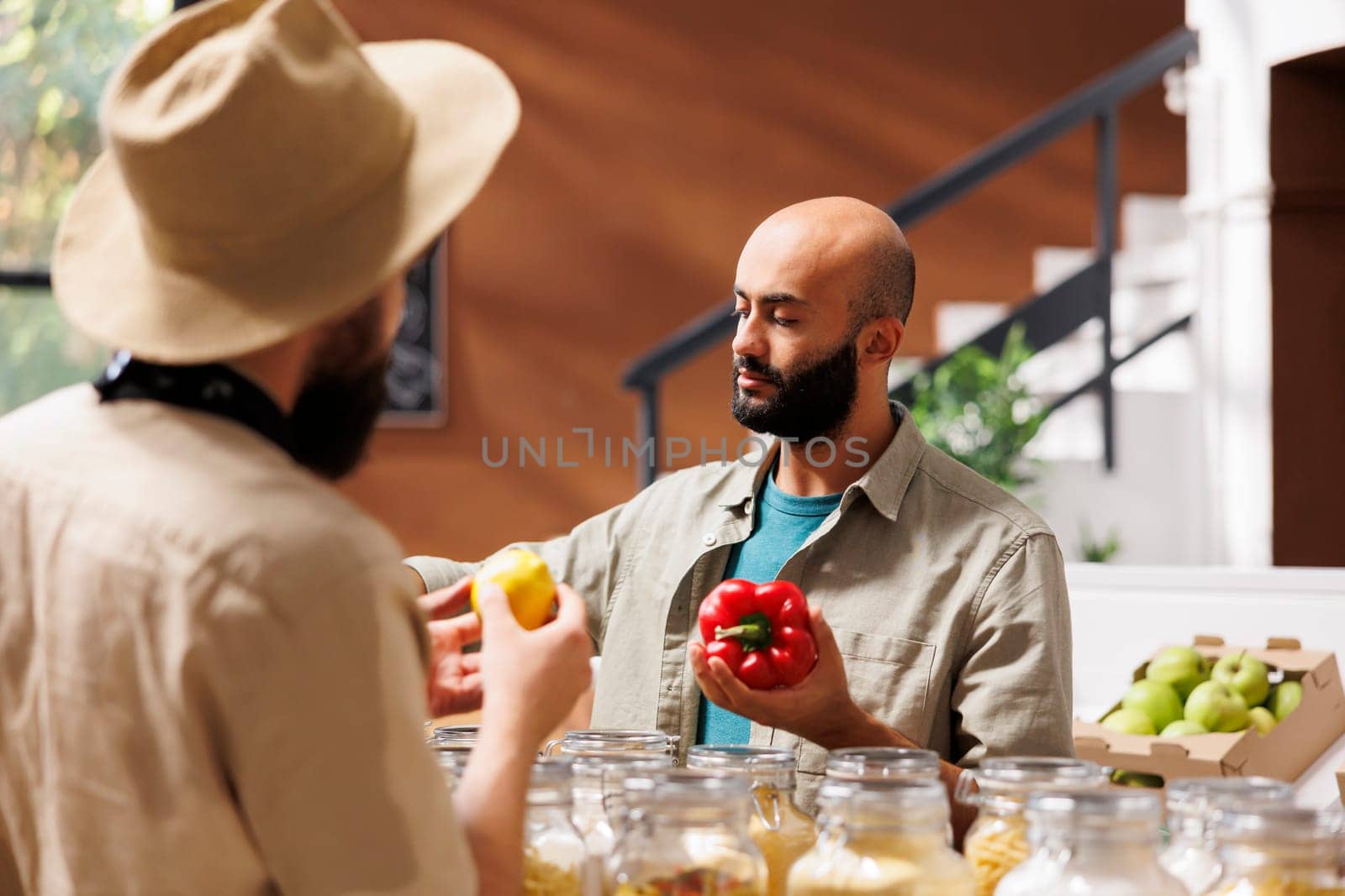 Middle eastern customer holds a red pepper while a male storekeeper looks at him while holding a lemon, in an eco-friendly grocery store. Salesman conversing with client on healthy lifestyle.