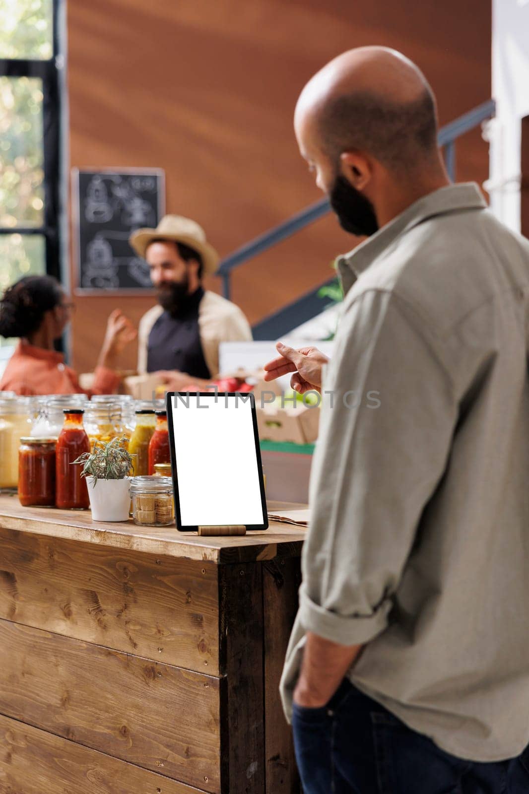 Middle Eastern man looking at electronic tablet with blank white screen placed on table filled with bulk products in reusable glass containers. Client using device with isolated mockup template.