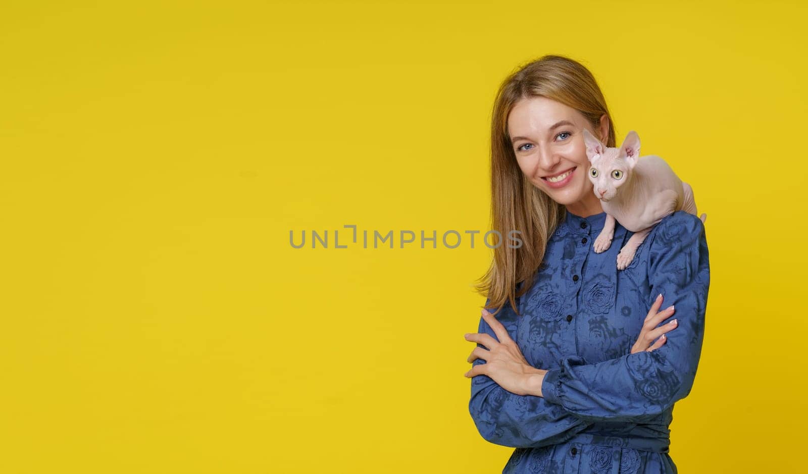 Smiling blond woman showcases unique bond with Egyptian cat Sphinx as it sits on shoulder against yellow background. Special companionship between woman and exotic feline friend, pet owner and cat. High quality photo