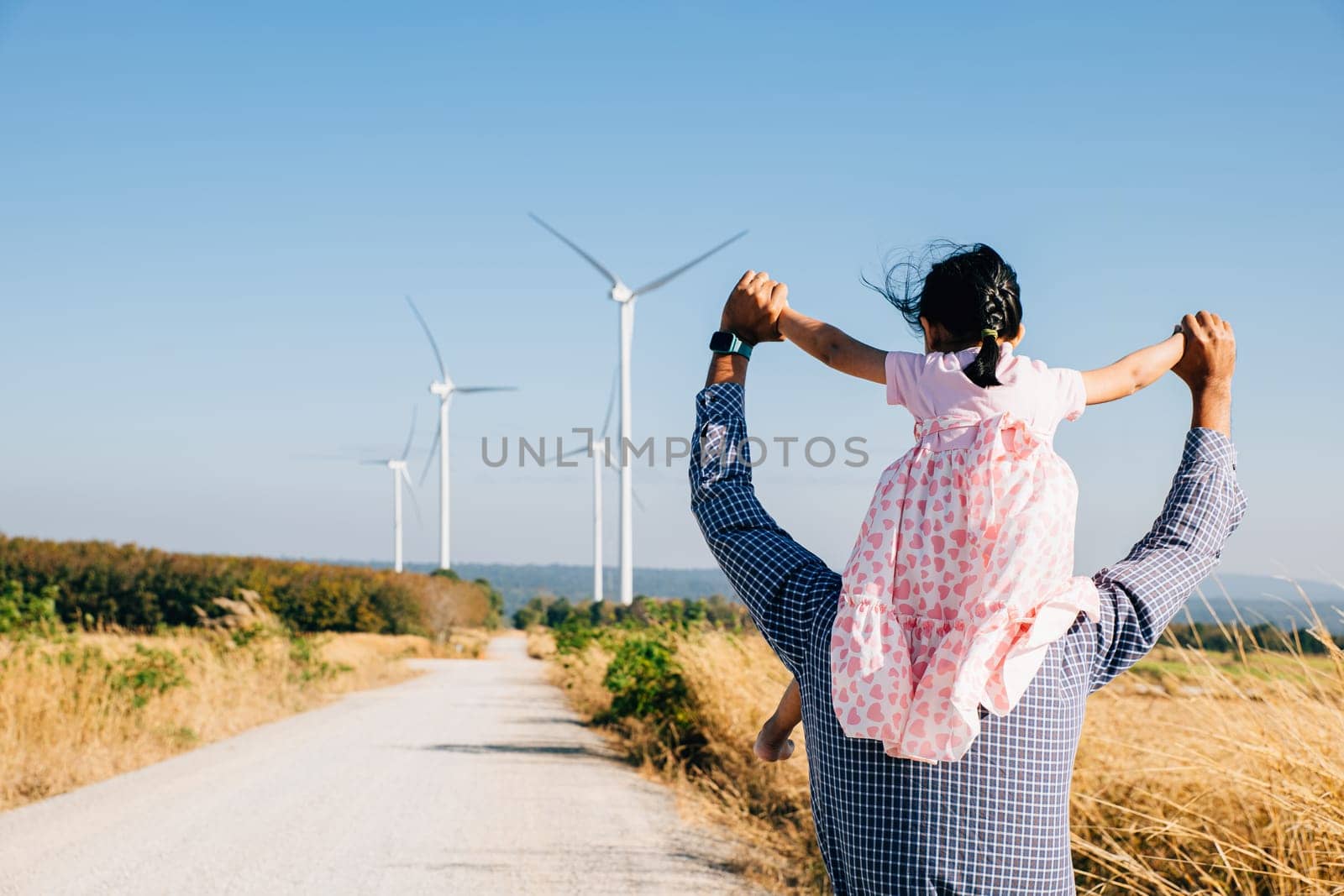 Father joyfully carries daughter exploring wind farm by Sorapop