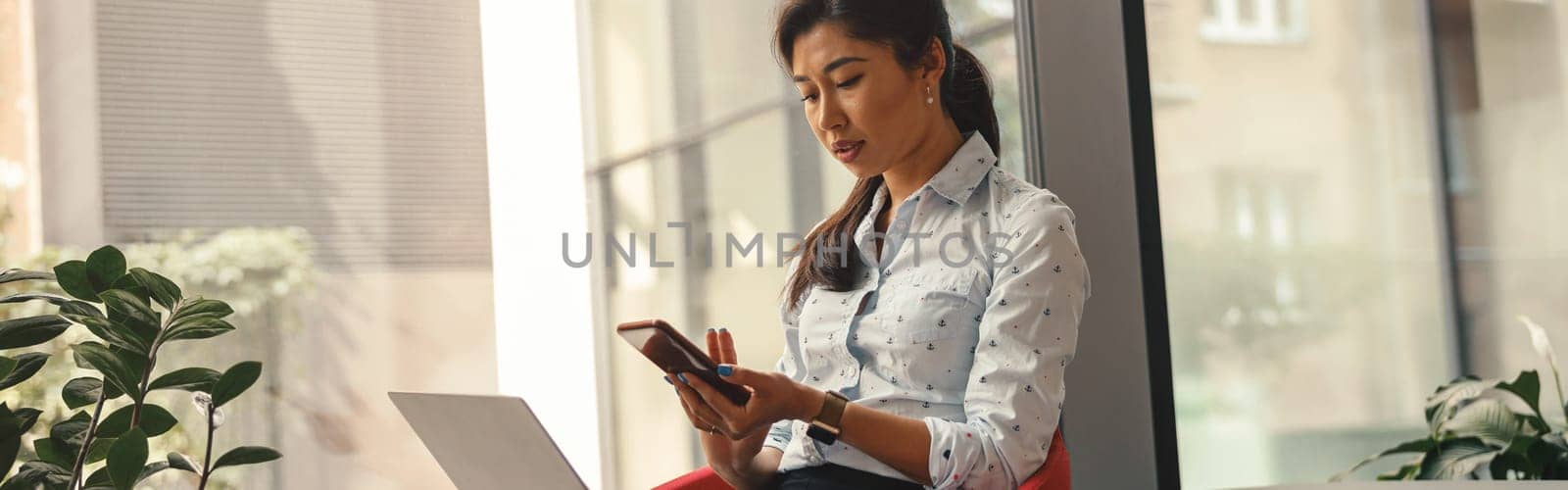Woman entrepreneur looking on phone while working on laptop in office