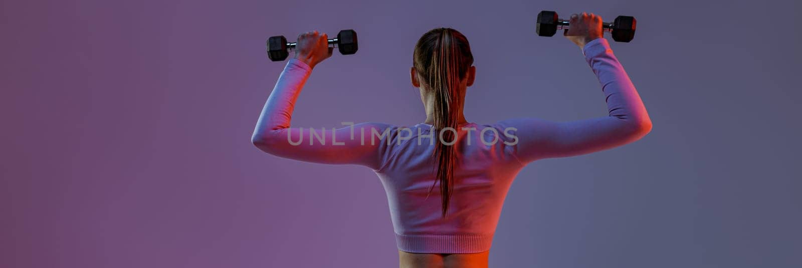 Back view of fitness woman doing exercises with dumbbells on studio background with color filter by Yaroslav_astakhov