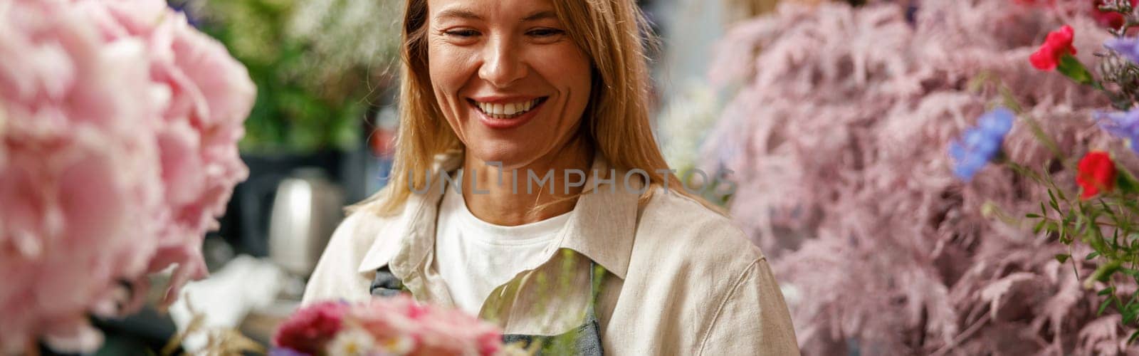 Smiling woman flower shop owner in apron looking on bouquet of flowers at florist store by Yaroslav_astakhov