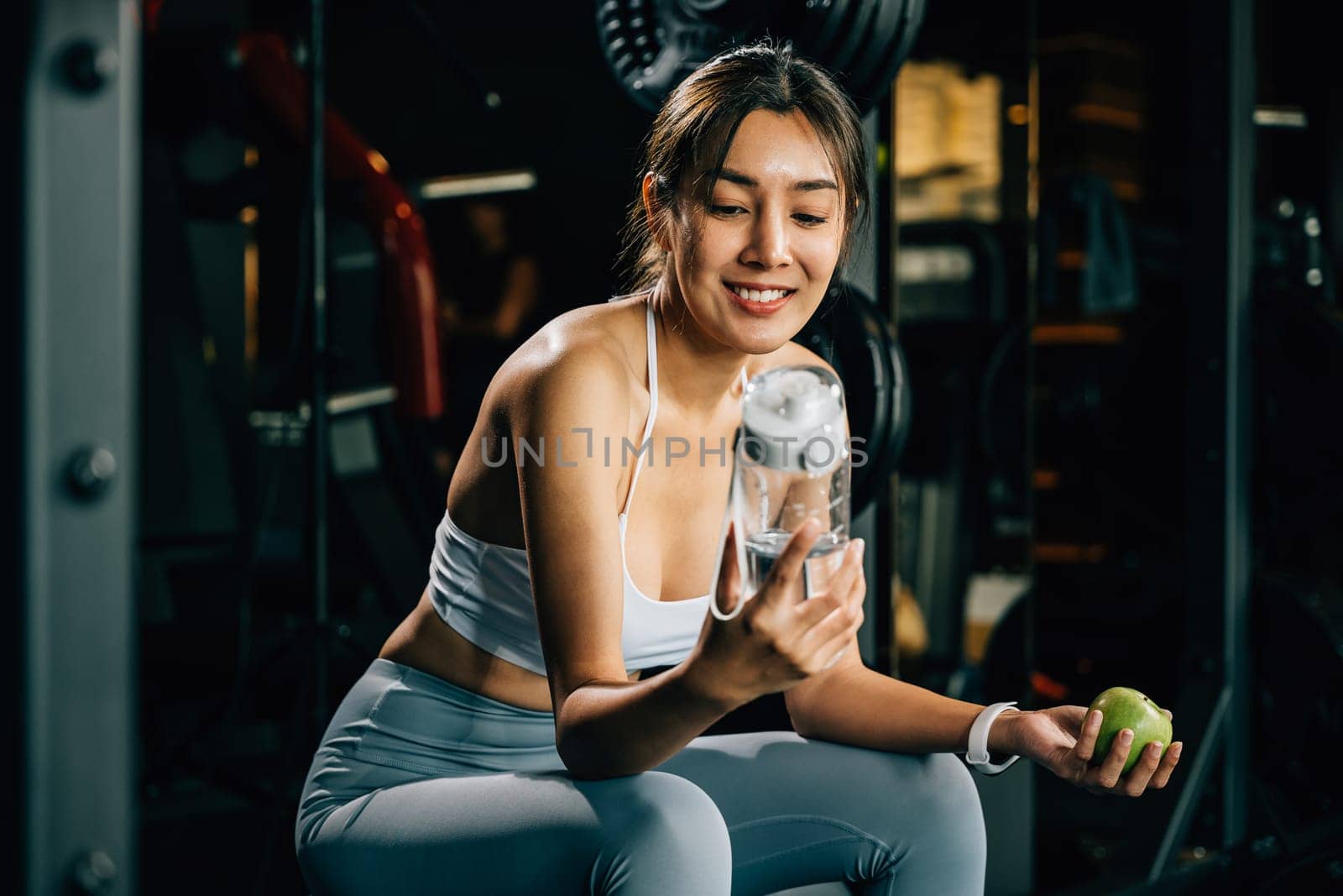 Asian woman holding a green apple and a water bottle in a fitness gym by Sorapop