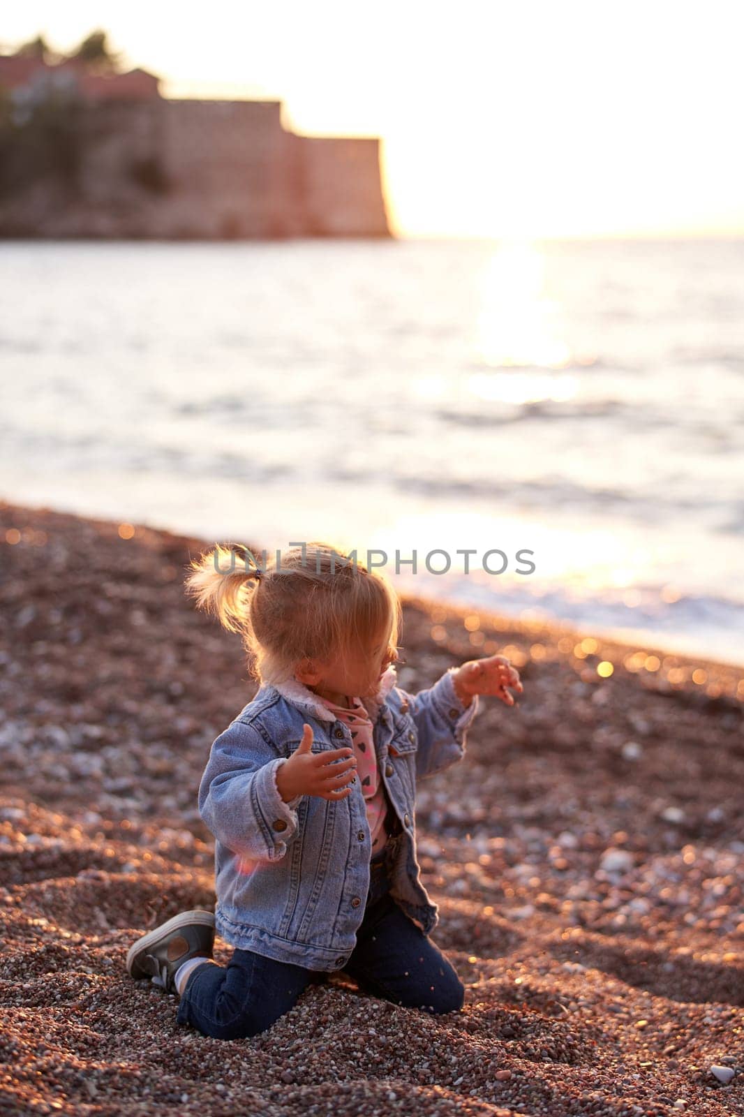 Little girl sitting on her knees on a pebbly beach with her hands up and fingers spread by Nadtochiy