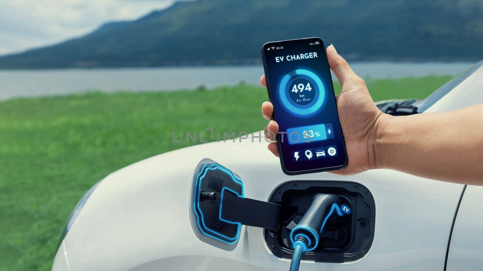 Businessman holding smartphone display battery status interface by smart EV mobile application while EV car recharging electricity from charging station with natural lake outdoor background.Peruse