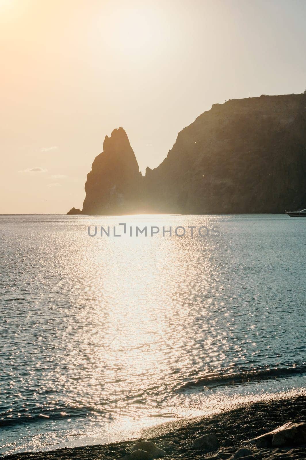 A red burning sunset with the silhouette of a cliff over the sea. Abstract nature summer or spring ocean sea background. Small waves on golden warm water surface with bokeh lights from sun.