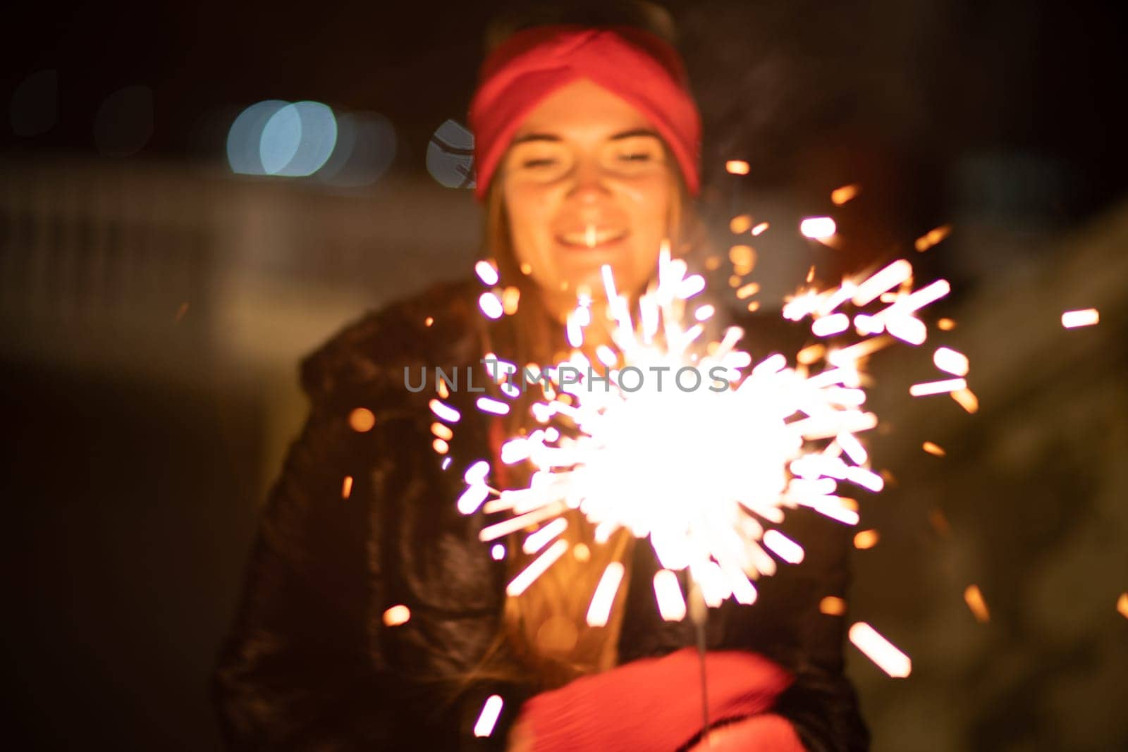 Woman holding sparkler night while celebrating Christmas outside. Dressed in a fur coat and a red headband. Blurred christmas decorations in the background. Selective focus.