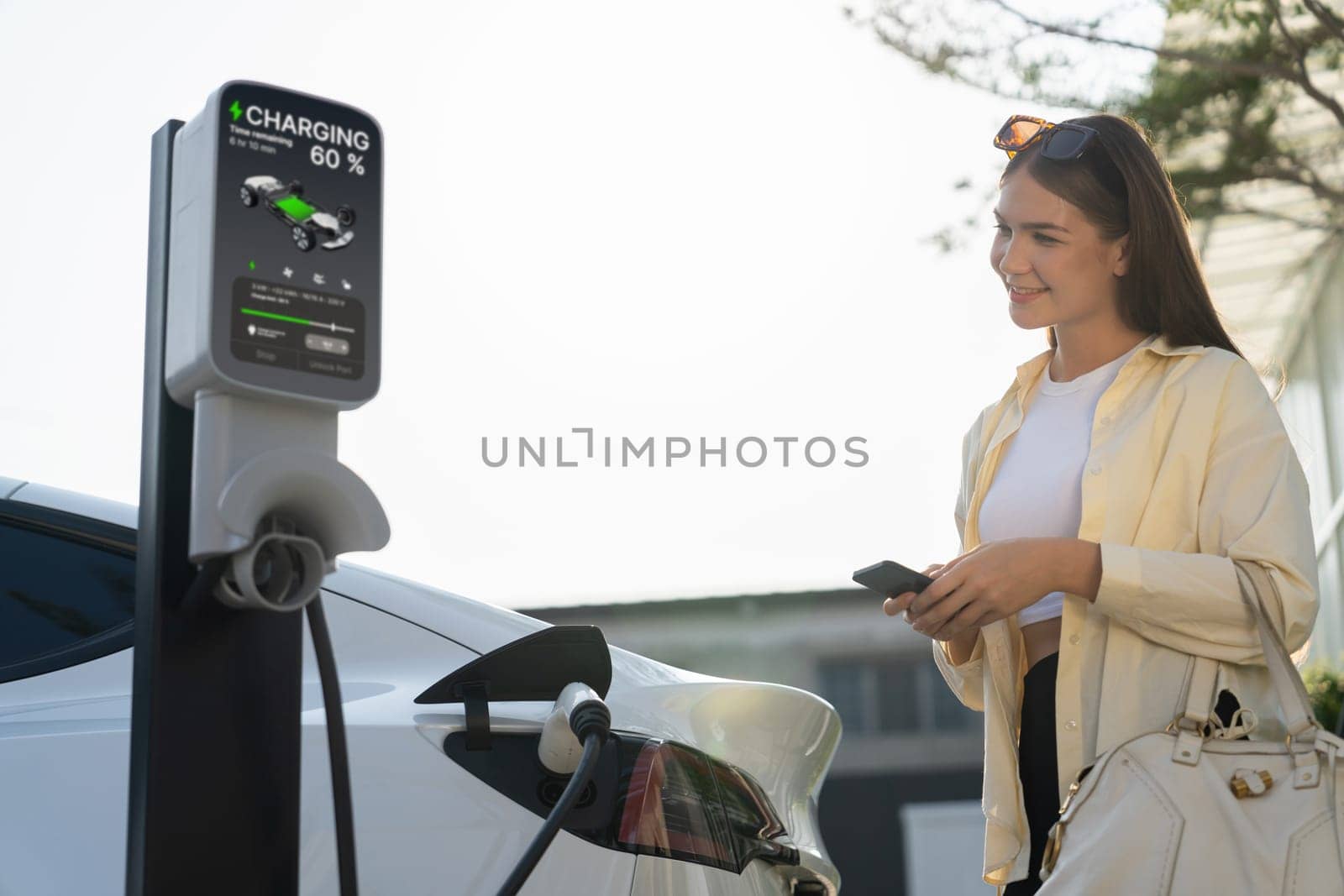 Young woman holding shopping bag and use smartphone to pay for electricity for recharging EV car battery from charging station at city mall parking lot. Modern woman go shopping by eco car. Expedient