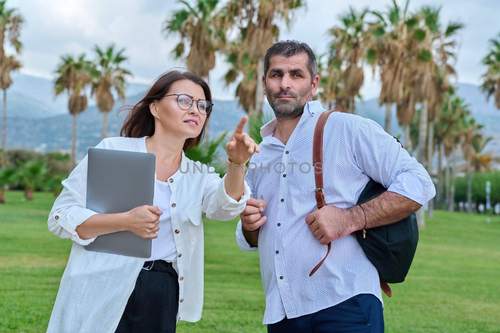 Middle aged business colleagues talking in the park. Mature man with backpack and woman with laptop meeting outdoors, positive smiling communicating