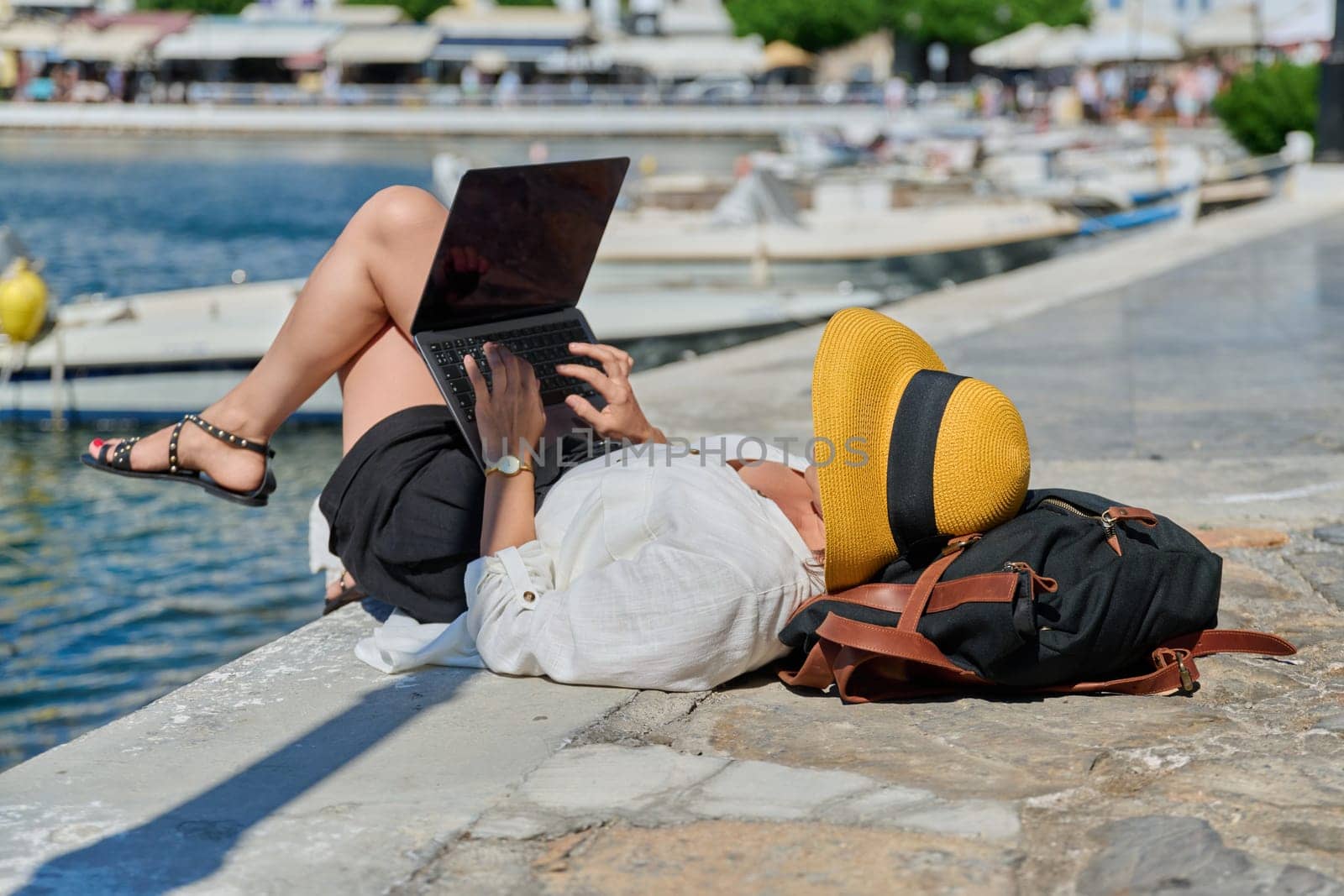 Woman in hat typing on laptop, water bay boat background. Remote work, freelance, tourism, travel, business and entrepreneurship, technology concept