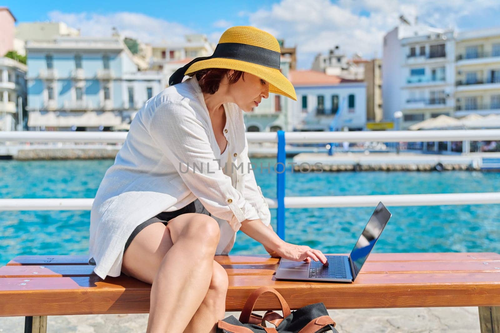Mature woman in hat with laptop on promenade in city. Female freelancer tourist traveler typing text on laptop, sea harbor old city background. Tourism, summer, technology, business, travel, freelance