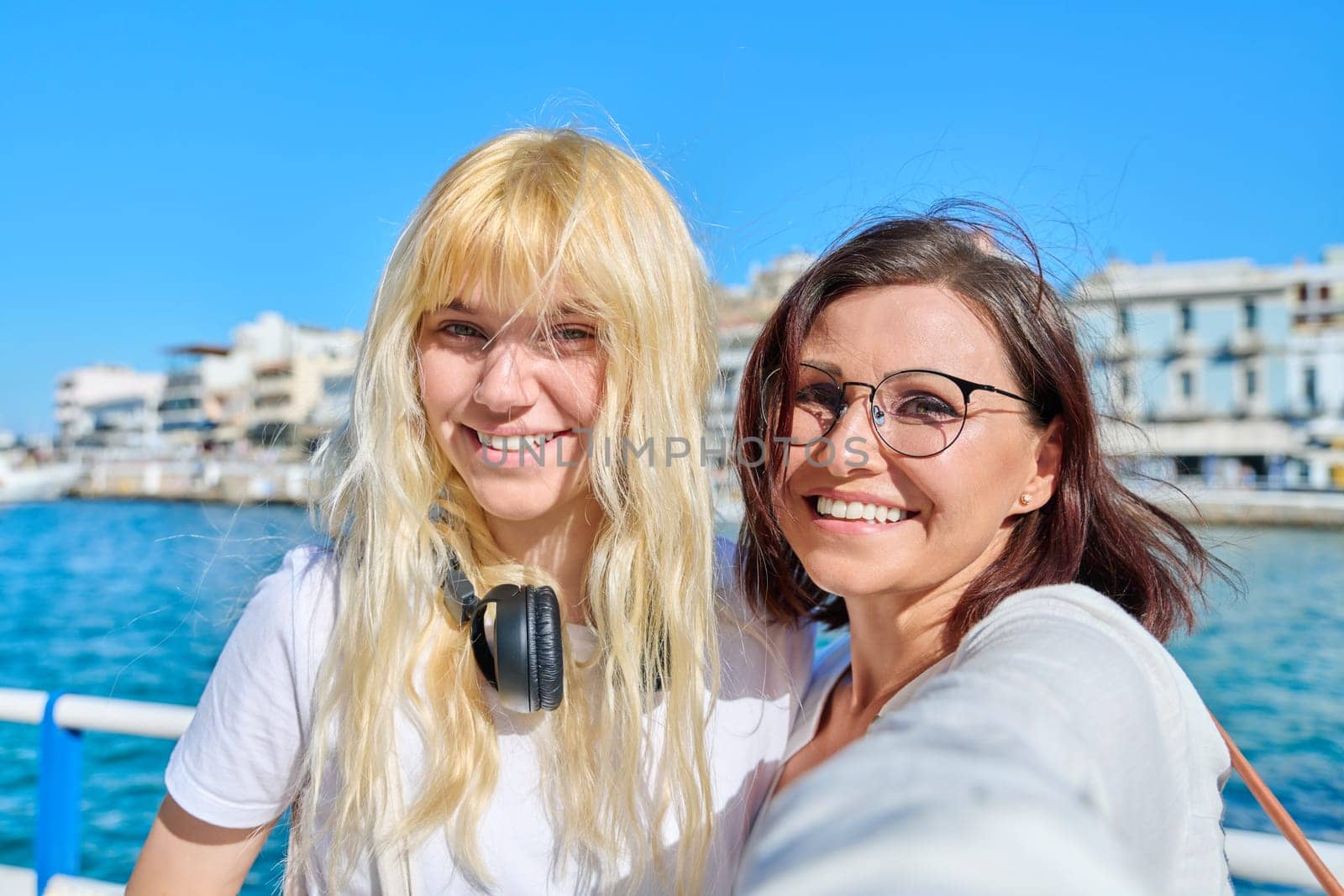 Portrait of happy smiling mom and teenage daughter looking at camera together, faces close-up. Sea bay in old tourist town, sunny summer day. Relationship parent teenager, lifestyle, family vacation