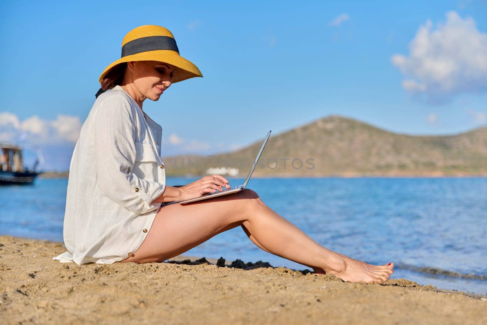 Mature woman in hat using laptop on the beach. Middle-aged female on vacation at the sea, working on a laptop. Remote work, freelance, business, nature, leisure, successful life, 40s people concept