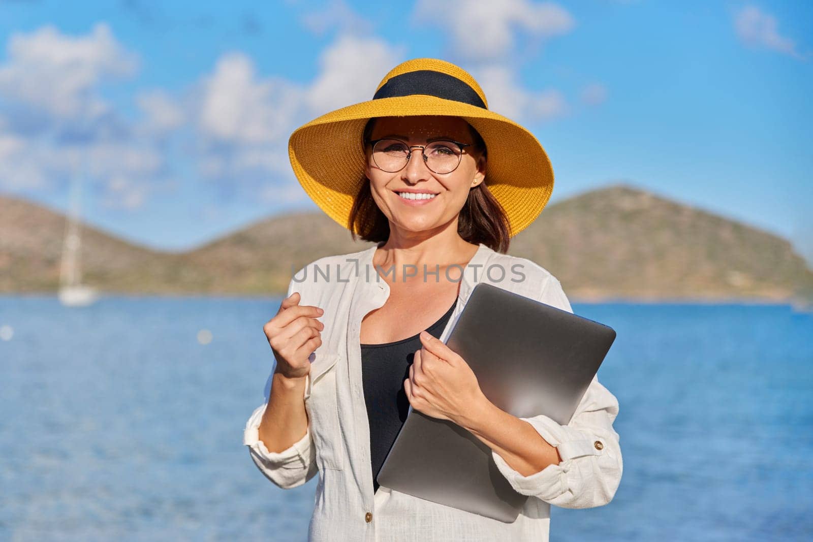 Portrait of happy mature woman in straw hat with laptop in hand on beach, looking at camera. Freelance, business, remote work, leisure, relaxation, tourism, technology, middle aged people