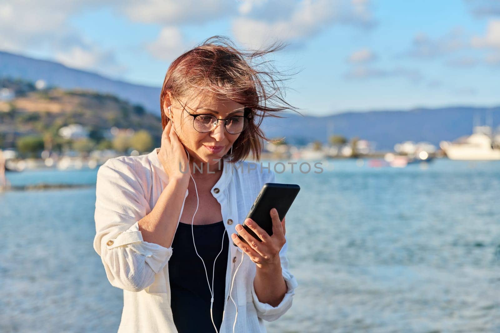 Middle-aged smiling woman relaxing on beach wearing headphones with smartphone. Mature female listening audio podcast, watching video. Leisure technology summer vacations, seaside resorts, people 40s