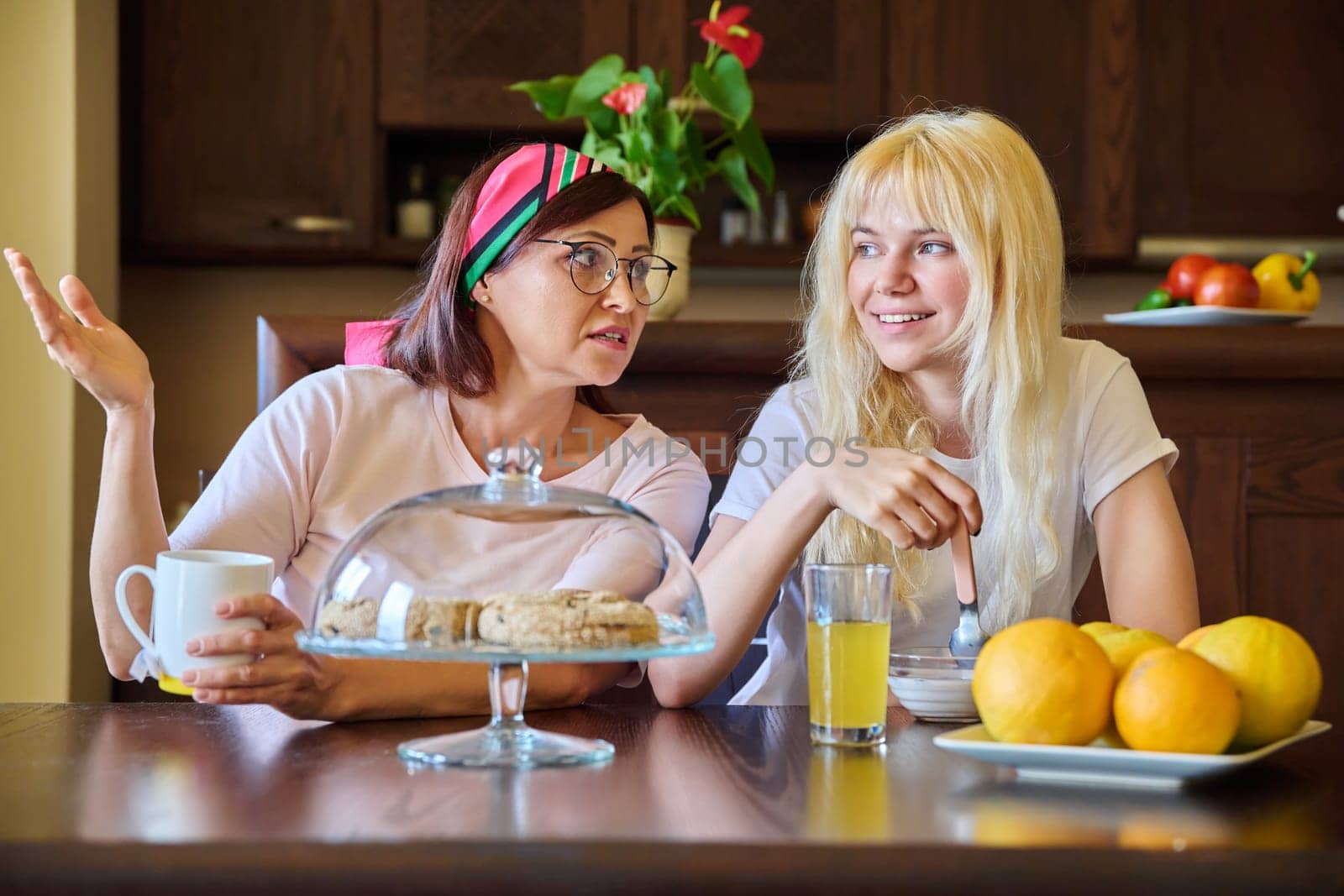 Mom and teenage daughter eating together, sitting at table at home in kitchen by VH-studio