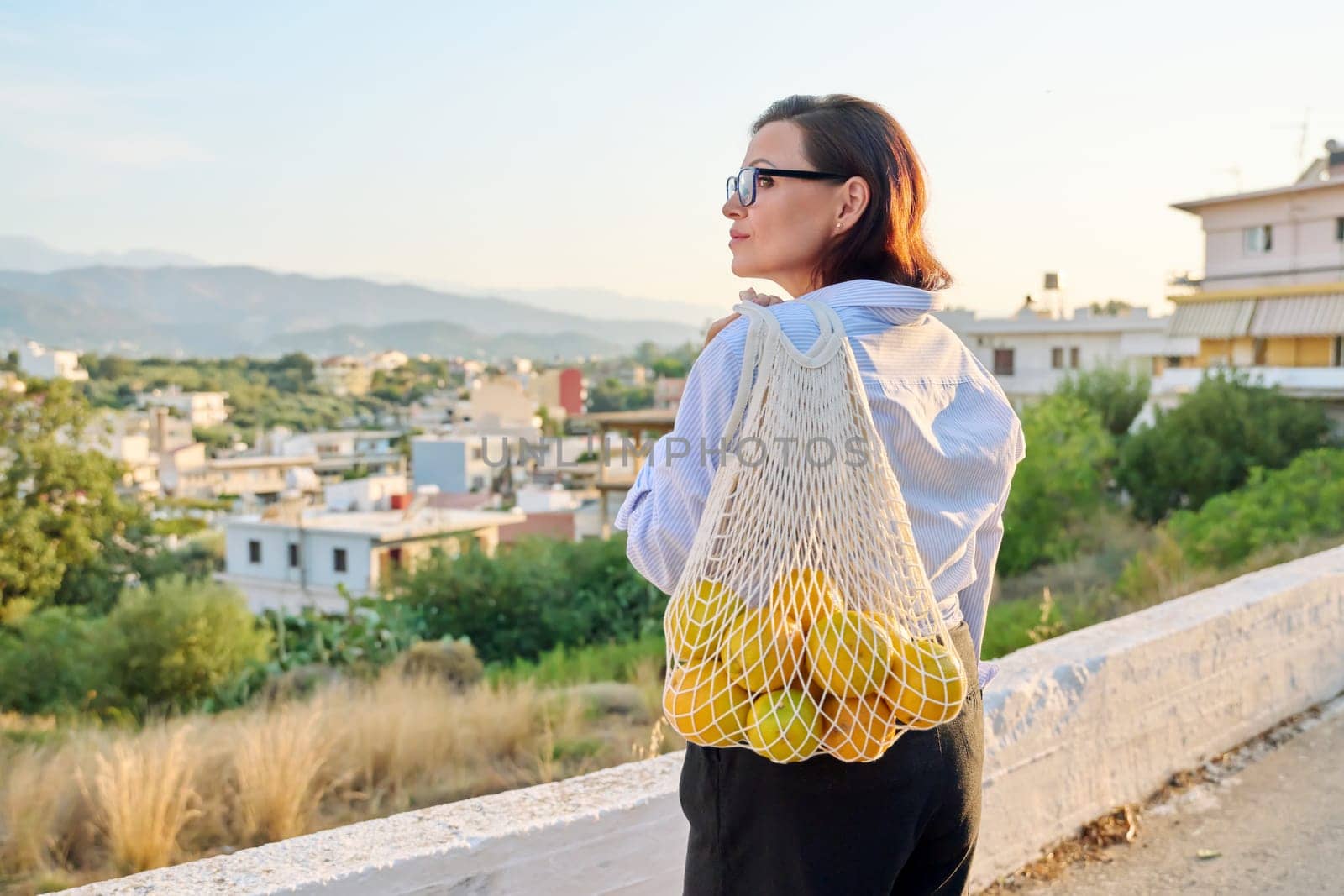 Eco tourism, woman walking relaxing in European mountain village, enjoying clean air, landscape, sunset, with a bag of fresh farm oranges. Nature, travel, ecology, lifestyle, people concept