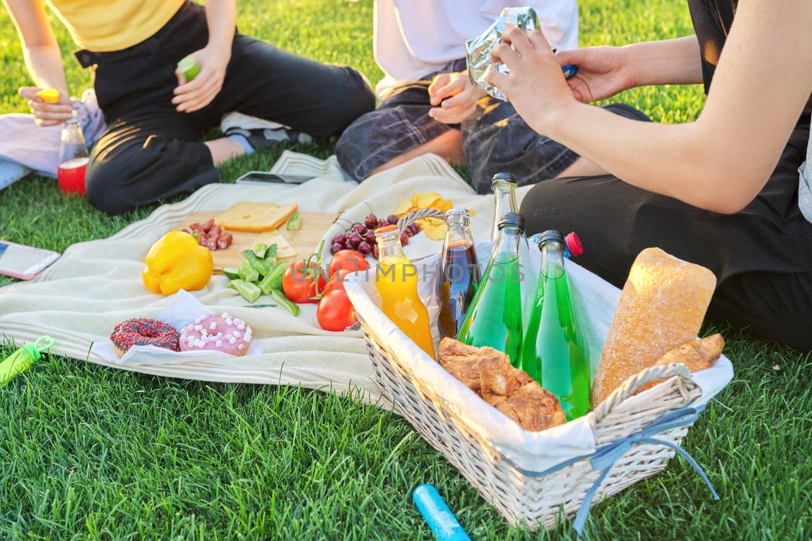 Close-up food and drink for picnic on green grass. Group of young people resting in nature, basket with bread, cheese fruits vegetables snacks juices on tablecloth. Youth, leisure, lifestyle, fun,
