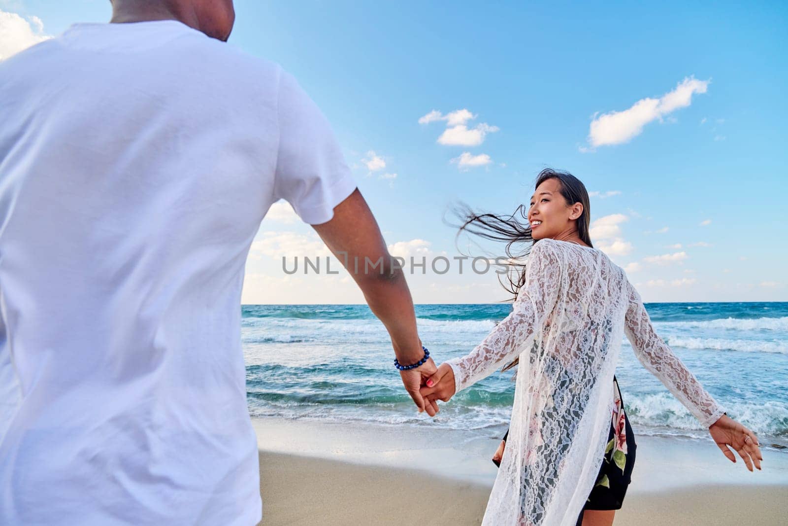 Loving happy couple walking holding hands on beach. Asian woman holding man's hand, sea sky background. Love, relationship, vacation, tourism, tourist travel to seaside resort, honeymoon concept