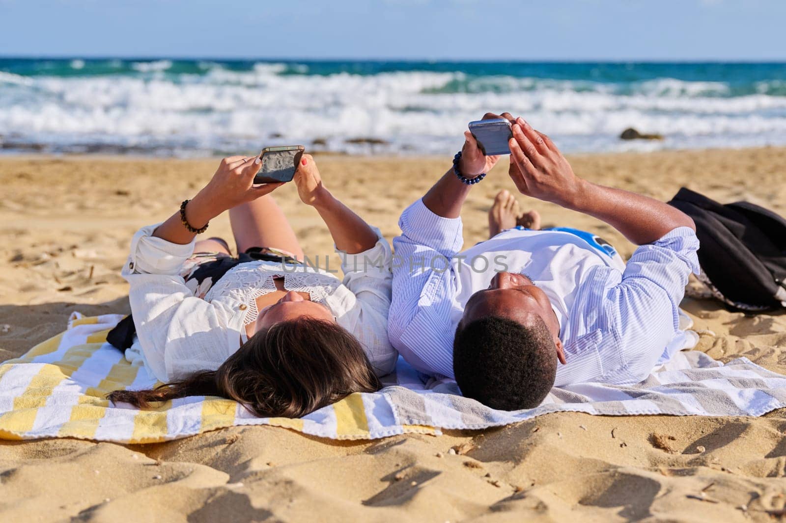 Young multicultural couple lying on beach using smartphones. Relaxing tourists lying on beach towels looking at smartphones. Lifestyle, technology, tourism, travel, nature, leisure, people concept