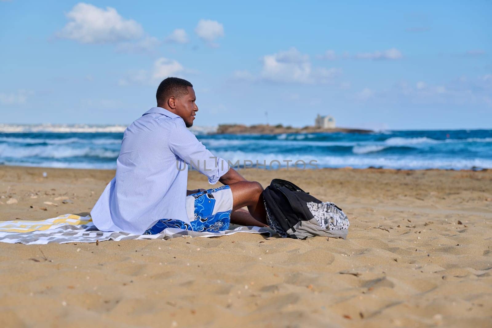 Young positive male tourist sitting resting on beach, copy space. Smiling African American male 30 years old sitting on sand, sea nature background. Lifestyle, travel, tourism, people concept