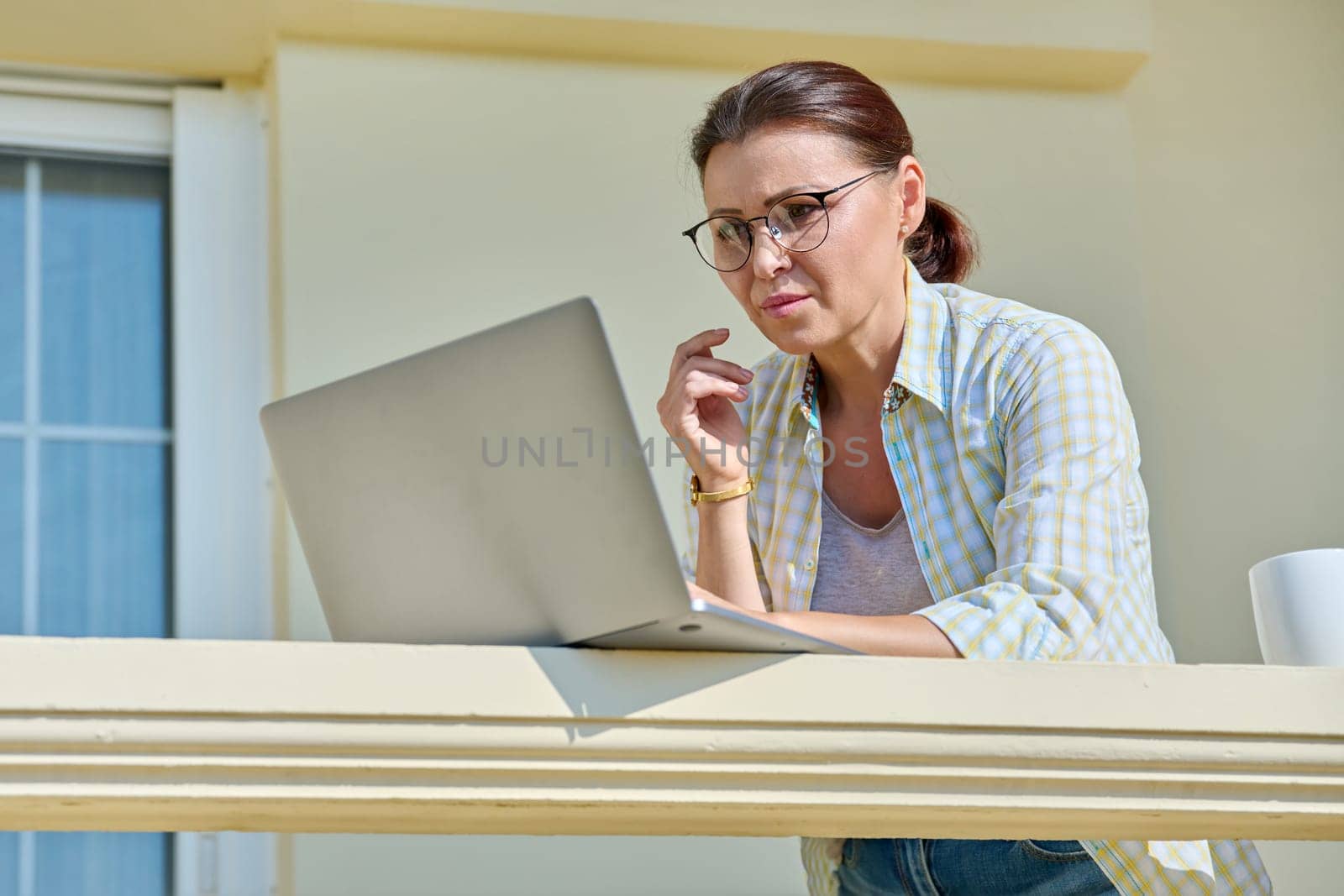 Serious focused middle aged woman with laptop on home balcony. Concentrated 40s mature business woman working remotely. Freelance, technology, work from home office, online training, blog, vlog
