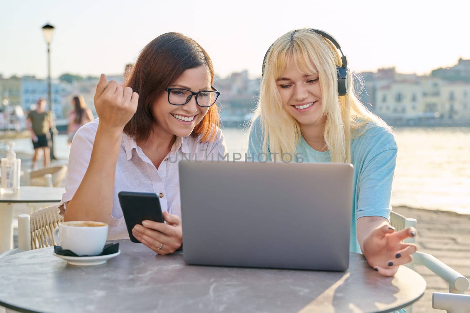 Mom and teenage daughter looking at laptop together, sitting in an outdoor cafe by VH-studio