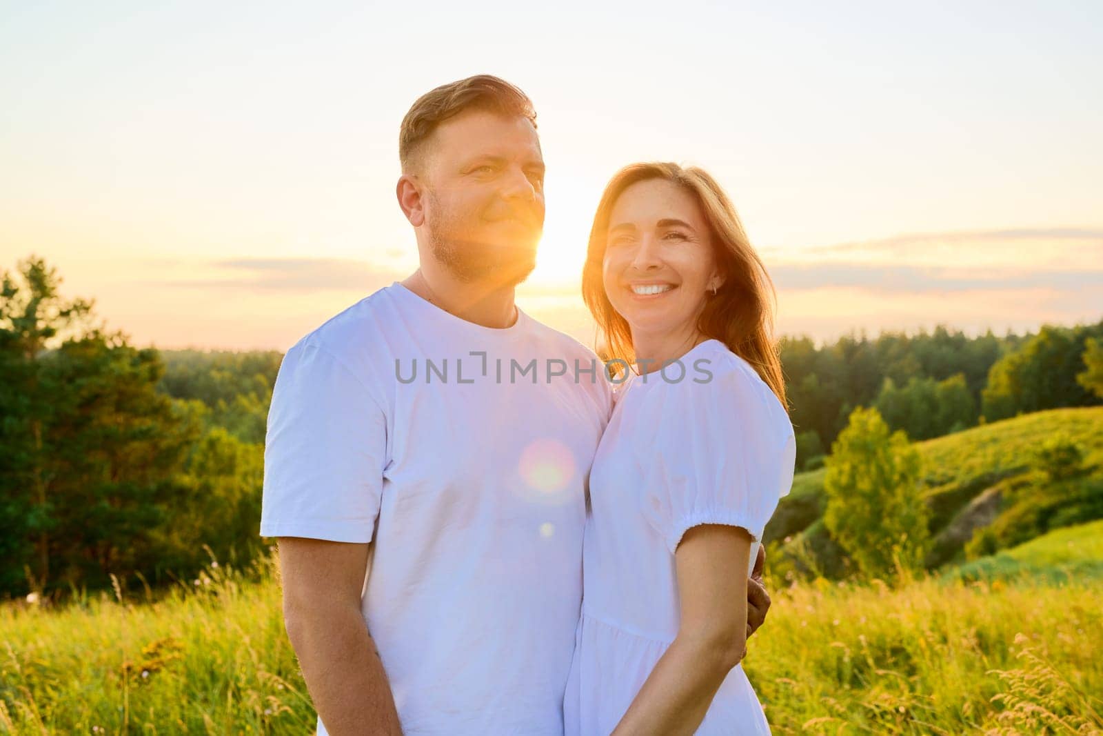 Outdoor portrait of adult happy loving hugging couple. Smiling 40s man and woman in white clothes, together at sunset, scenic natural landscape background. Relationship, love, happiness, family people