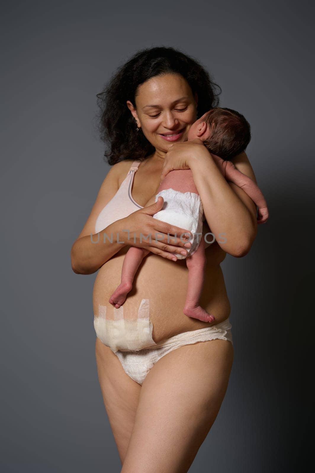 Happy mother in lingerie, showing naked belly with bandage hiding scars after c-section cesarean and postpartum stretch marks, smiling while holding her newborn baby, isolated gray studio background