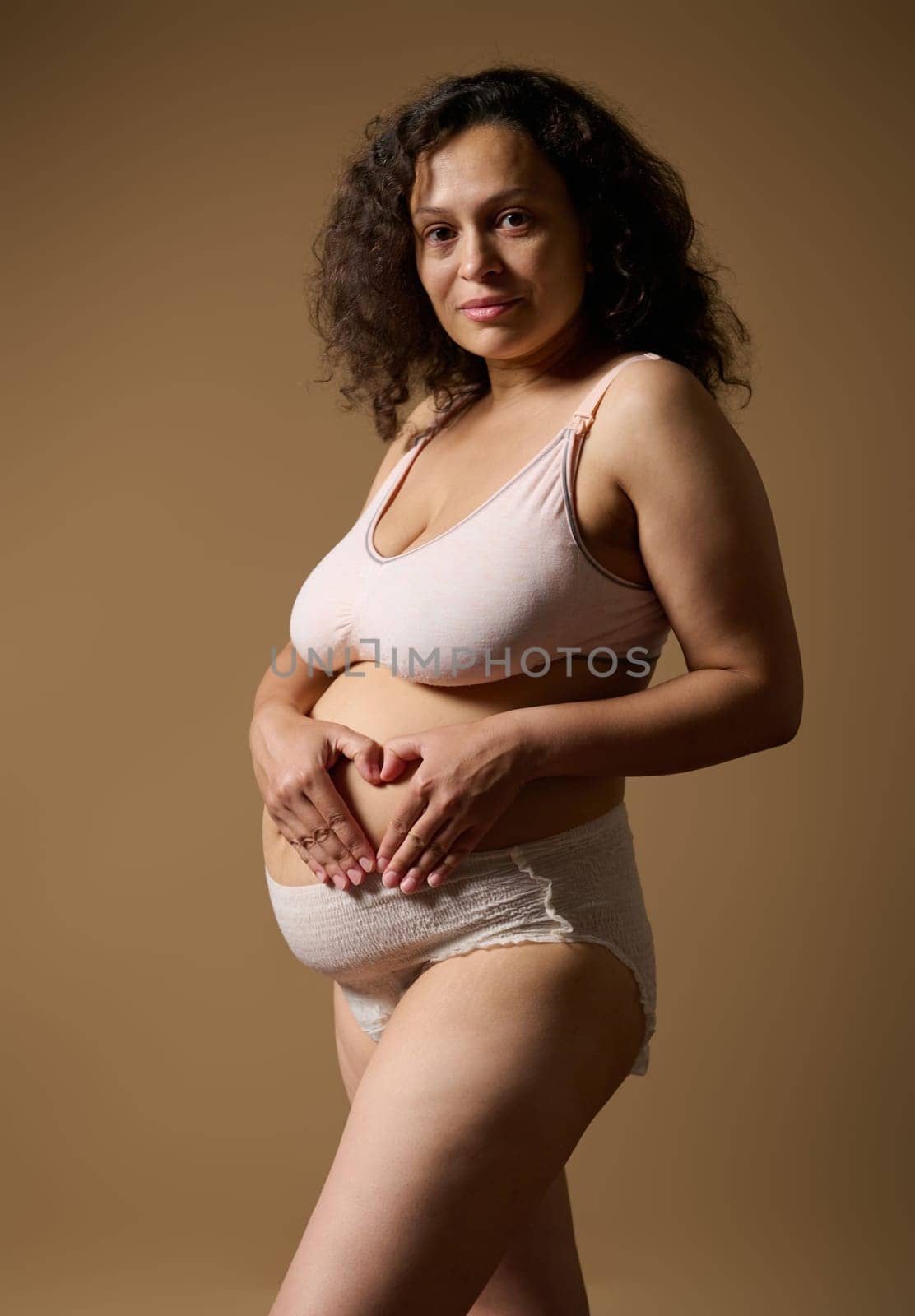 Confident woman in underwear, making heart shape over her postnatal belly, expressing confidence looking at camera. Mother with visible postpartum body marks, isolated on beige background