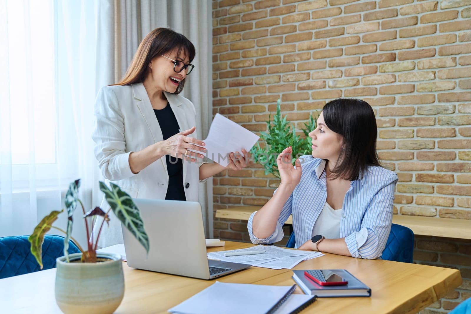 Two joyful happy smiling business women colleagues sitting at large table in office with business papers contracts. Business ceo work law finance mentoring consulting teamwork people job concept