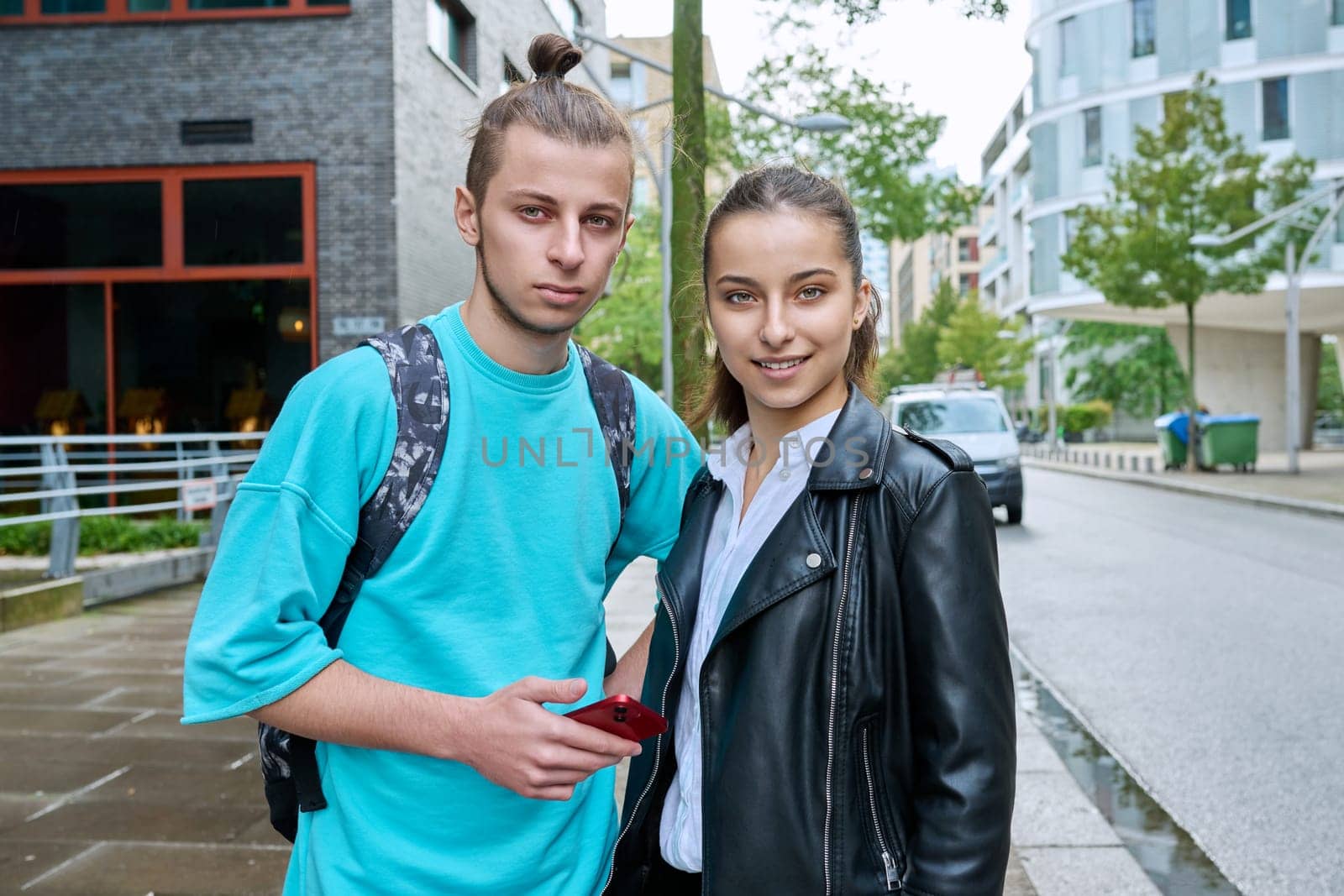 Outdoor portrait of teenagers students friends smiling guy and girl with smartphones looking at camera on street of modern city. College, 17-19 years old, urban lifestyle, adolescence youth concept