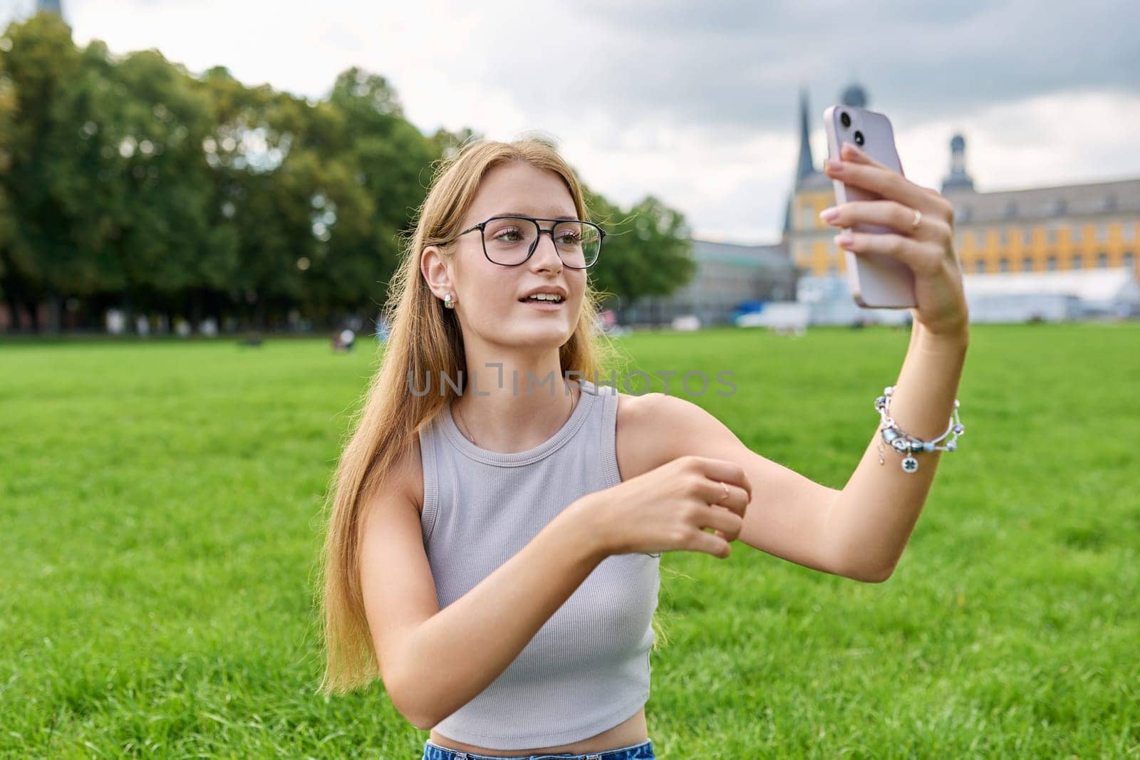 Teenage girl student having video call on smartphone, resting sitting on grass lawn near university building. Technology, leisure, communication, youth