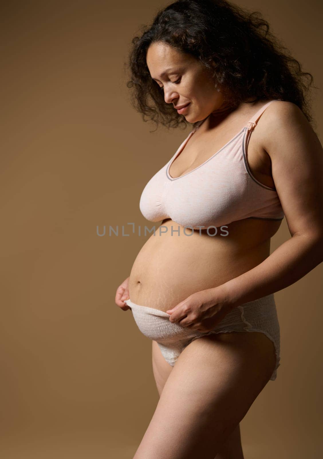 Confident authentic pretty woman mother in underwear, posing with her postnatal naked belly with flaws and stretch marks few days after childbirth, isolated beige studio backdrop. Postpartum recovery