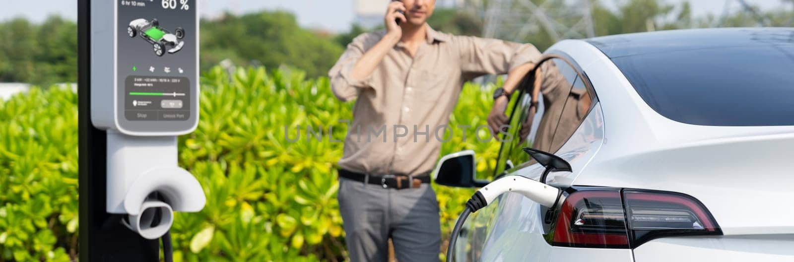 Man talking on the phone while recharge EV car battery at charging station connected to power grid tower electrical as electrical industry for eco friendly car utilization. Panorama Expedient